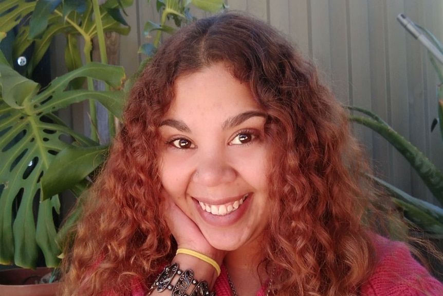 A smiling young woman with long curly hair sits in front of some plants.