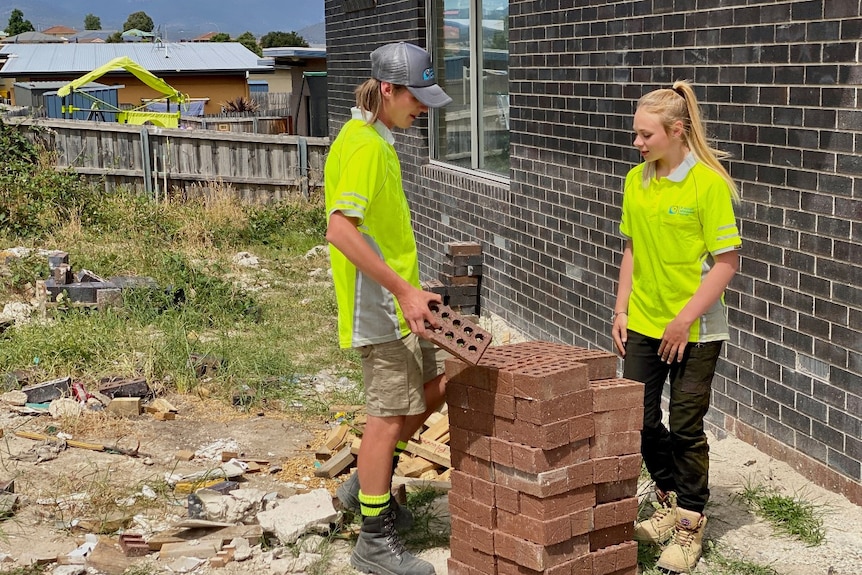 A teenage boy and teenage girl chat while moving a pile of bricks.