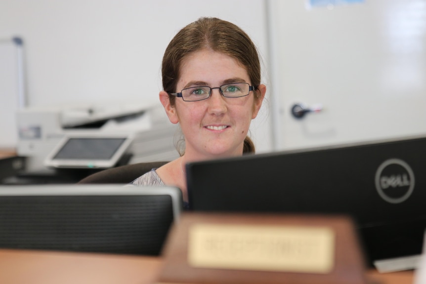 A woman wearing glasses looking over a pair of computer monitors. 