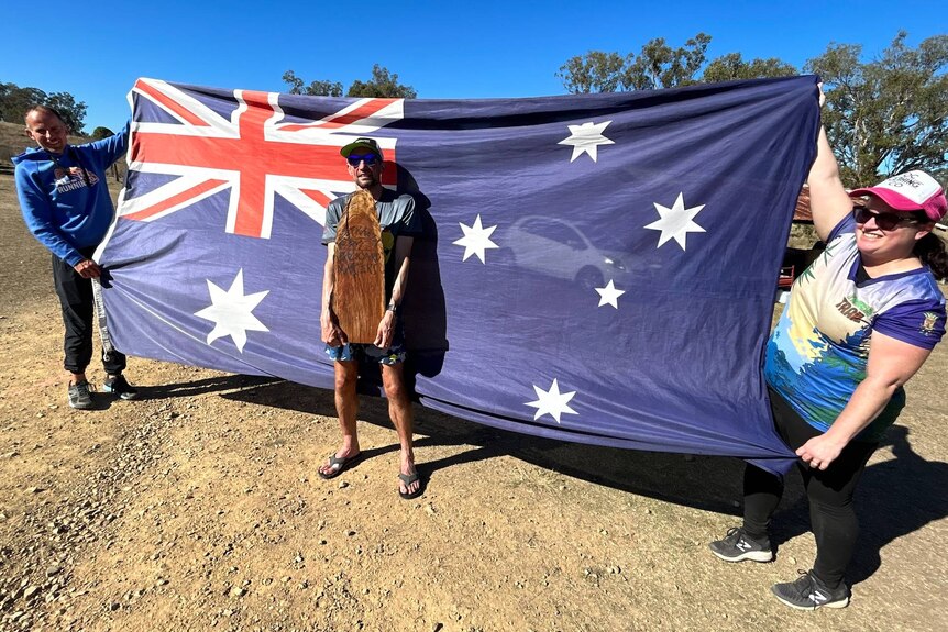 A man holding a large timber trophy while another man and a woman hold up a big Australian flag behind him.