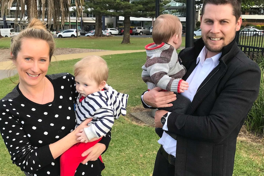 Mother and father of pre-term twins standing in a park holding a baby each smiling to camera