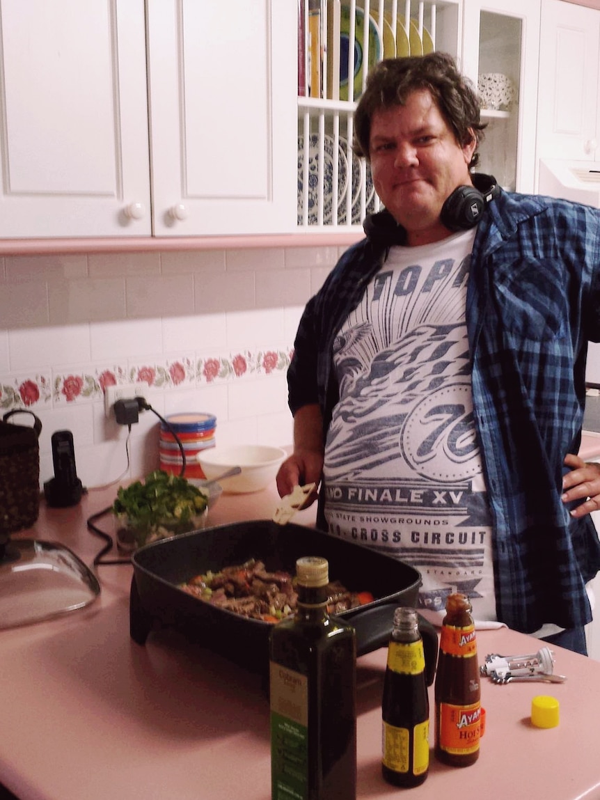 Man with headphones wearing a t-shirt and plaid buttoned shirt standing in a kitchen.