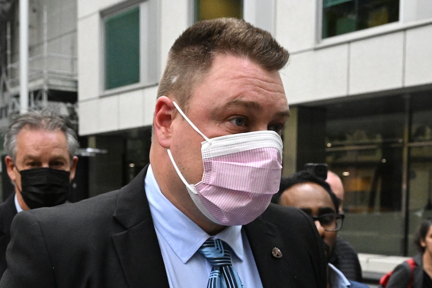 Brett Johnson, wearing a suit and face mask, leaves court surrounded by media.
