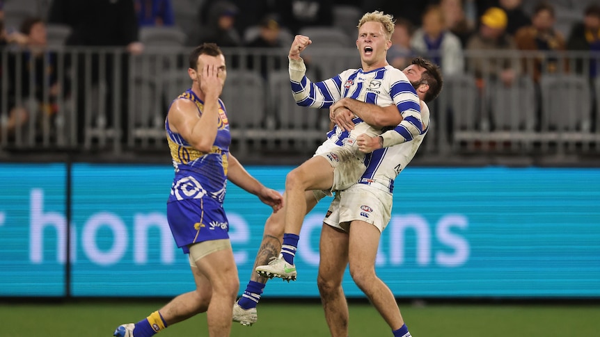 North Melbourne Kangaroos player Jaidyn Stephenson lifted by a teammate as he fist pumps. West Coast's Luke Shuey walks behind.