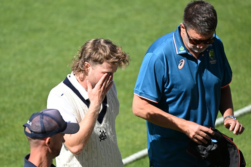 A concussed cricketer puts his hand to his face as he walks off the ground with medical staff.