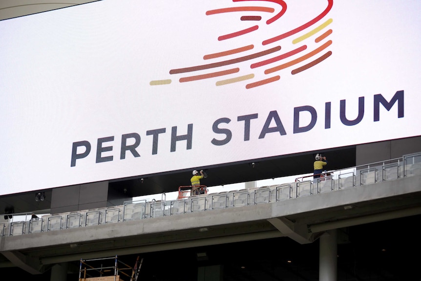 Workmen in high viz and hard hat put up Perth Stadium sign, inside the new stadium.