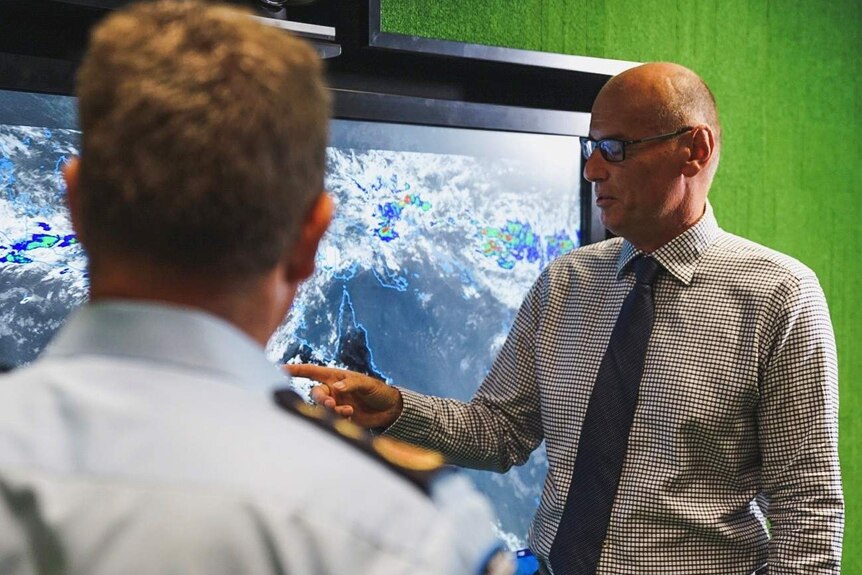 A man in a checked shirt and tie points at a large screen showing a weather system moving across Australia.