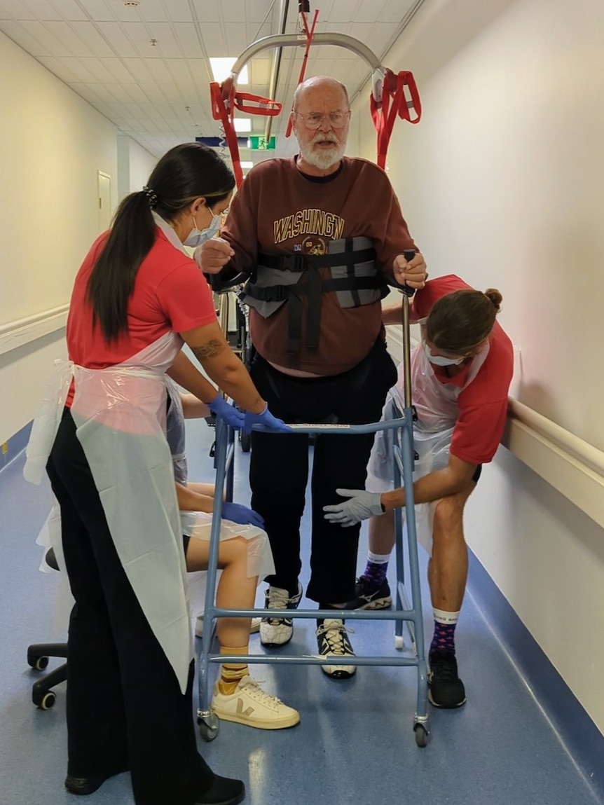 Man in a frame walking up a corridor with the help of nurses. 