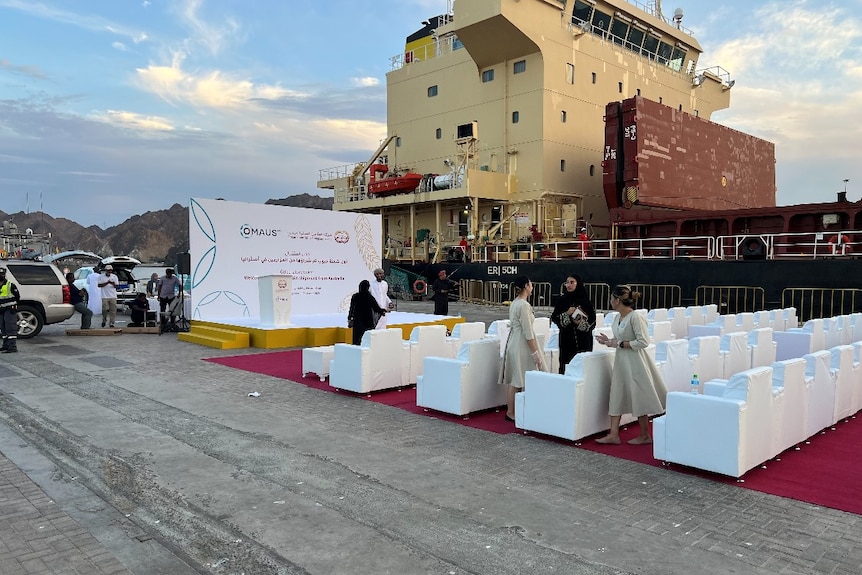 Lines of plush chairs on a red carpet at a dock in front of a cargo ship.