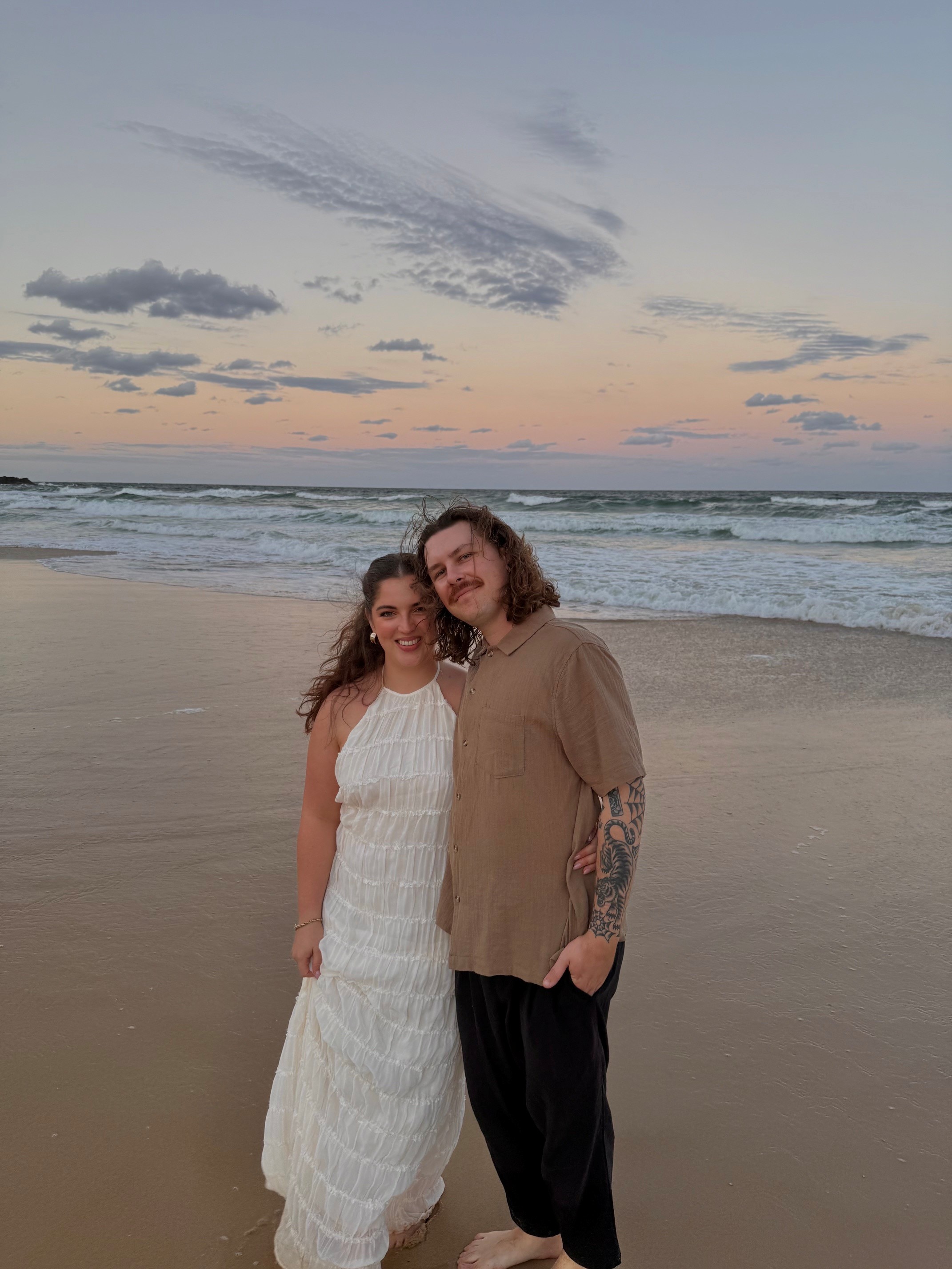 Woman in a white dress smiling at a man in a beige shirt on the beach at sunset