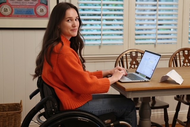 A young woman wearing an orange jumper sitting at a table using a computer.