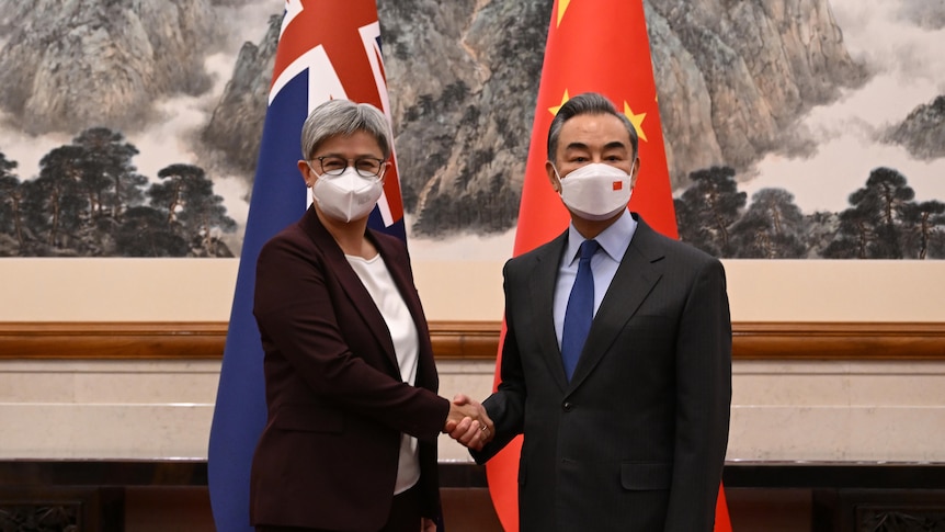Penny Wong and Wang Yi shake hands as they look at the camera, both in face masks. 