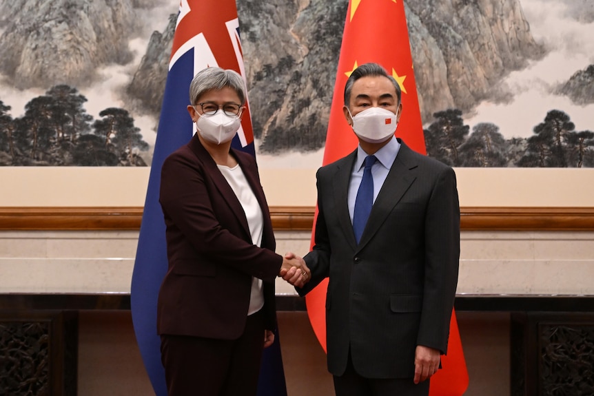 Penny Wong and Wang Yi shake hands as they look at the camera, both in face masks. 