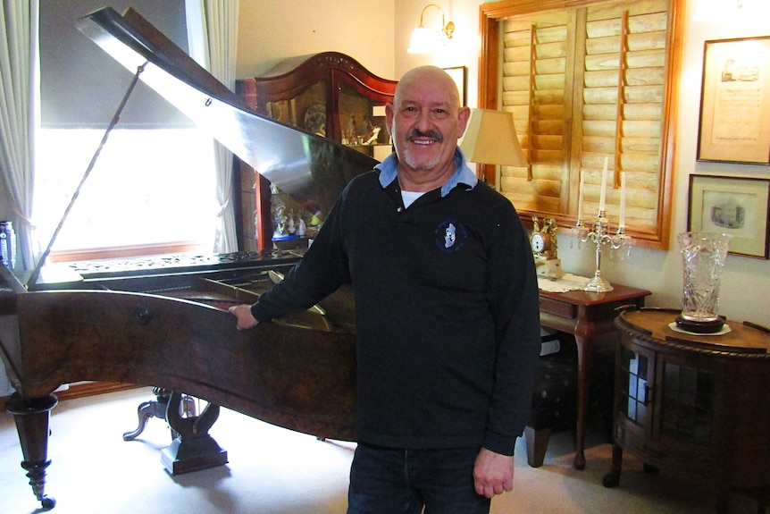 A bald man with a moustache stands in front of a grand piano