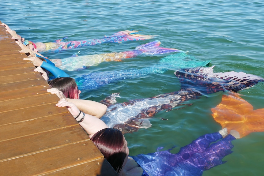 Five mermaids stretch their tails out while holding onto a boardwalk