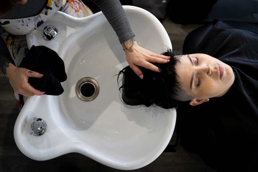 A person with their eyes closed lies in a salon chair with their hair in a sink behind them.