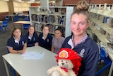 A female student stands with a teddy in the foreground with four female students sitting in the background in a school library.