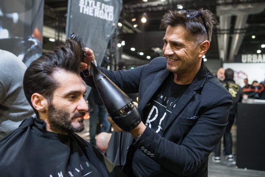 A man styles the hair of another man at an industry trade show.