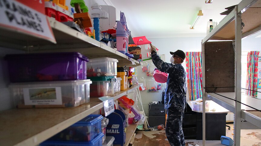 RAAF personnel wearing protective masks and gloves remove toys from shelves inside a library