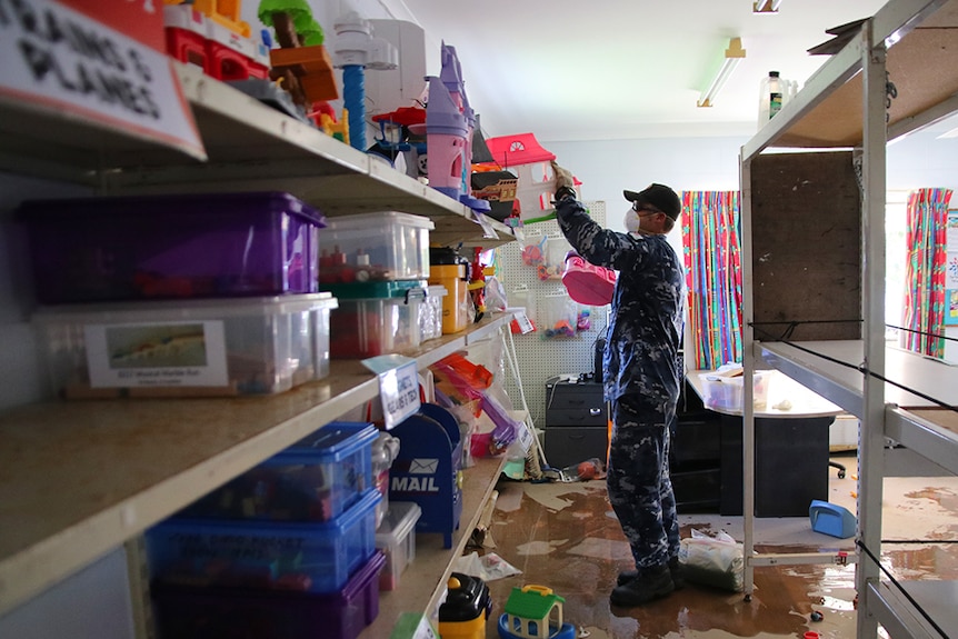 RAAF personnel wearing protective masks and gloves remove toys from shelves inside a library