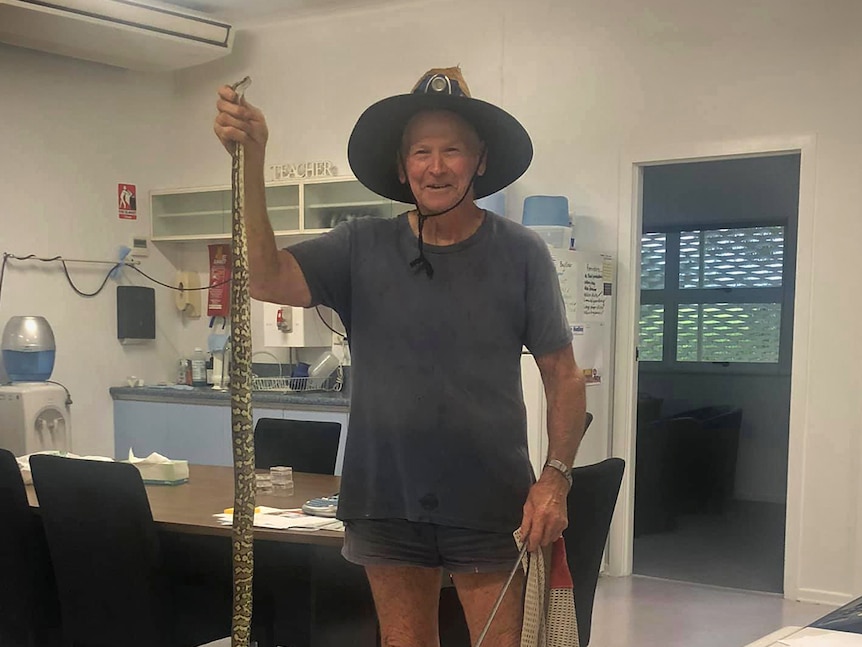 A snake catcher holds up a python, he is standing in a school office.
