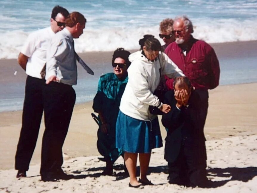 A group of people at a beach, with one man on his knees sobbing.