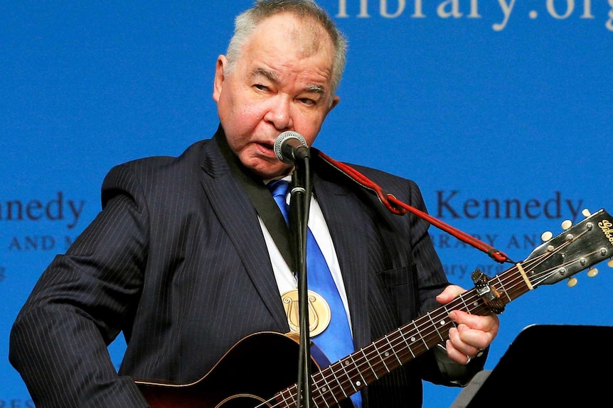 John Prine plays a guitar on stage in front of a microphone