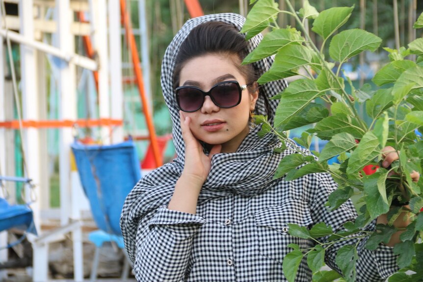 A young woman in a gingham shirt, large sunglasses and black head scarf 