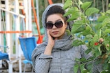 A young woman in a gingham shirt, large sunglasses and black head scarf 