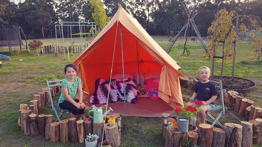 two kids sit in a camp set up in their back yard with an orange tent