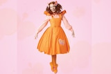 Emma Watkins jumps in an orange dress and shoes on a pink background.
