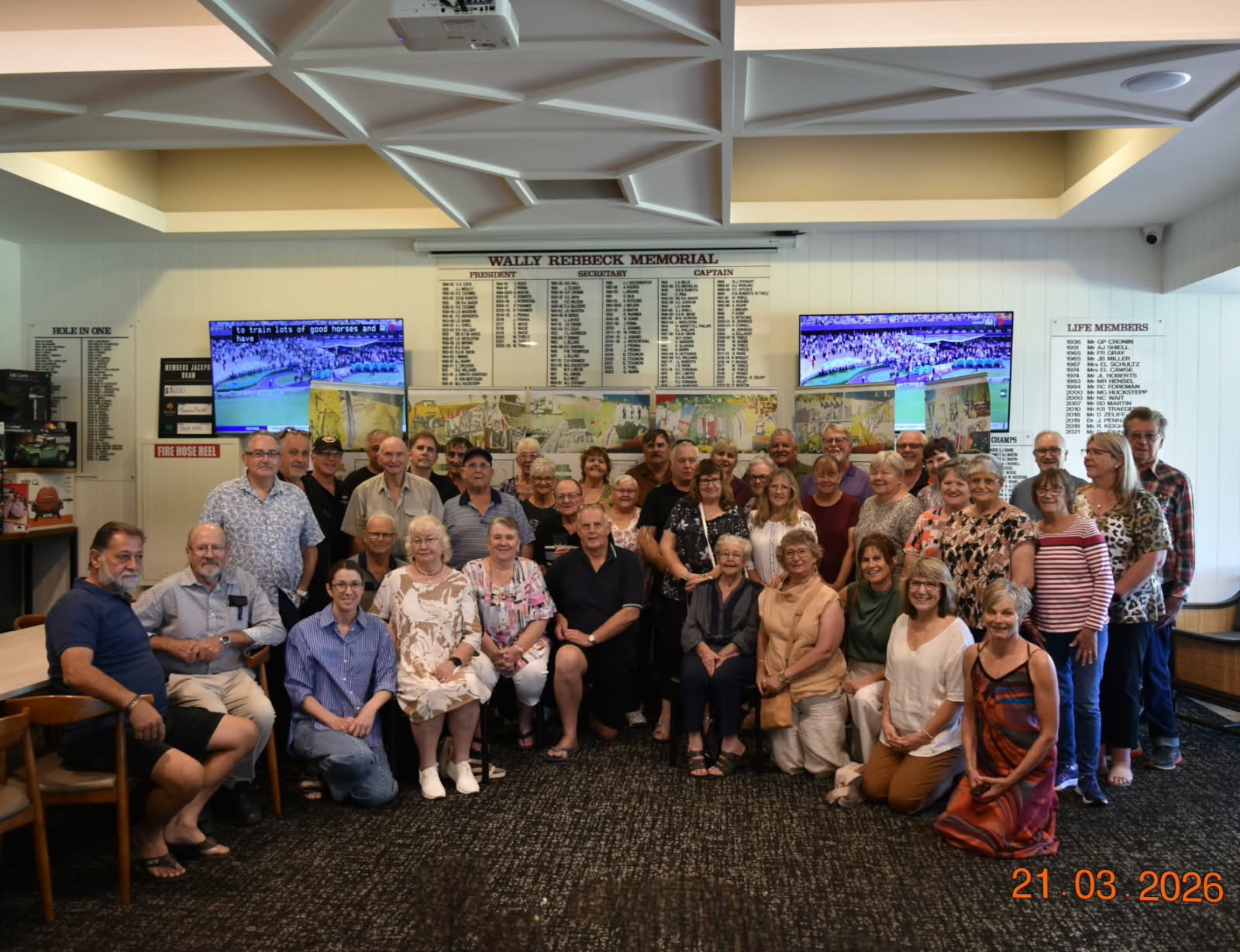 About 70 people stand and sit smiling in front of a banner at a golf club.