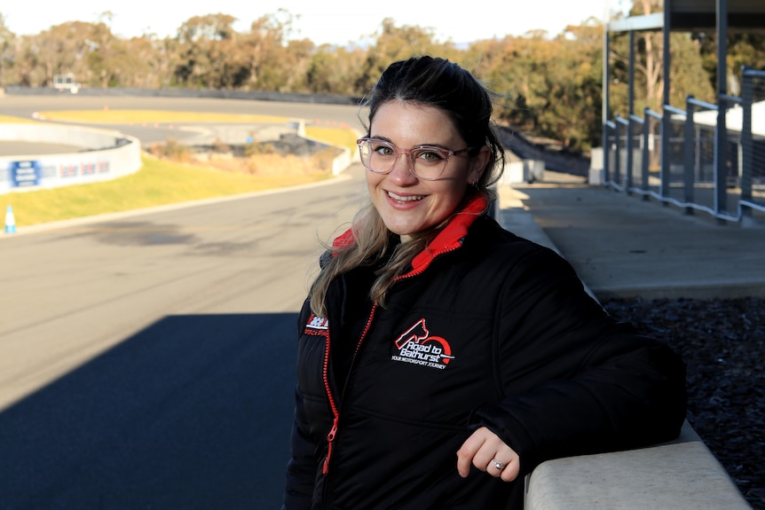Kinsey stands on the racing track wearing a black and red jacket and glasses, smiling.