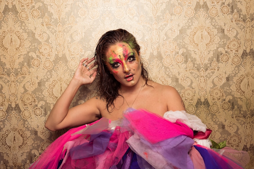 White woman with wet brown hair and pink, gold and green face paint sits in a pile of pink tulle fabric against a gold wall.