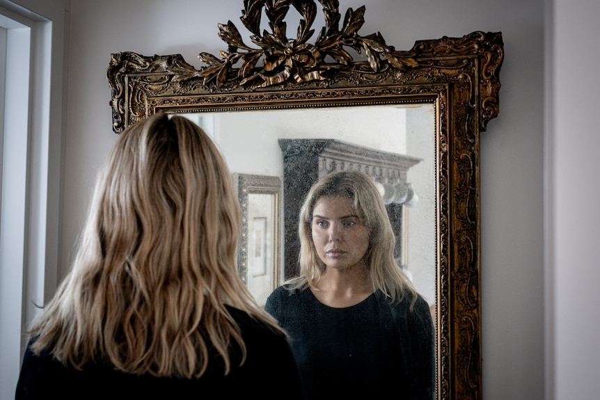 A young blonde woman looks at herself in a mirror.
