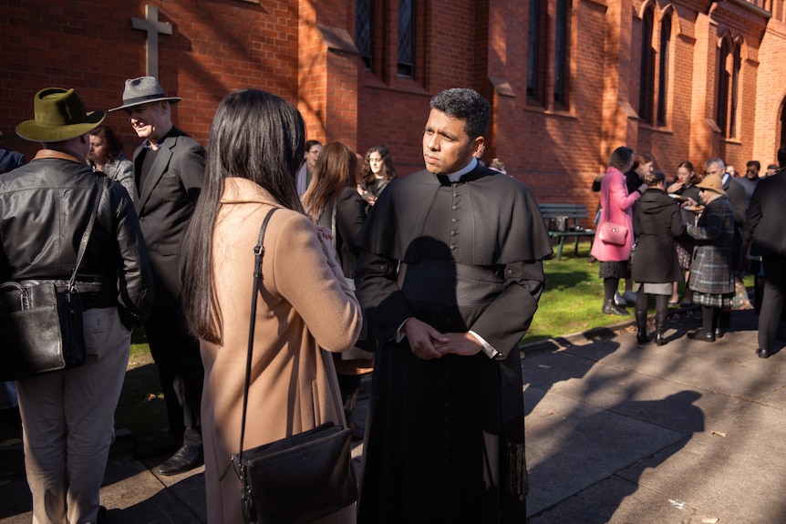 a priest in a black robe listens to a young woman outside a church