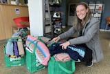 A woman with long brown hair, wearing a grey coat and black pants, smiles and crouches next to bags filled with essential items