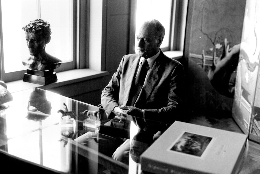 A man in a smart suit and tie sits at a glass desk with a sculpture on it. He looks pensive