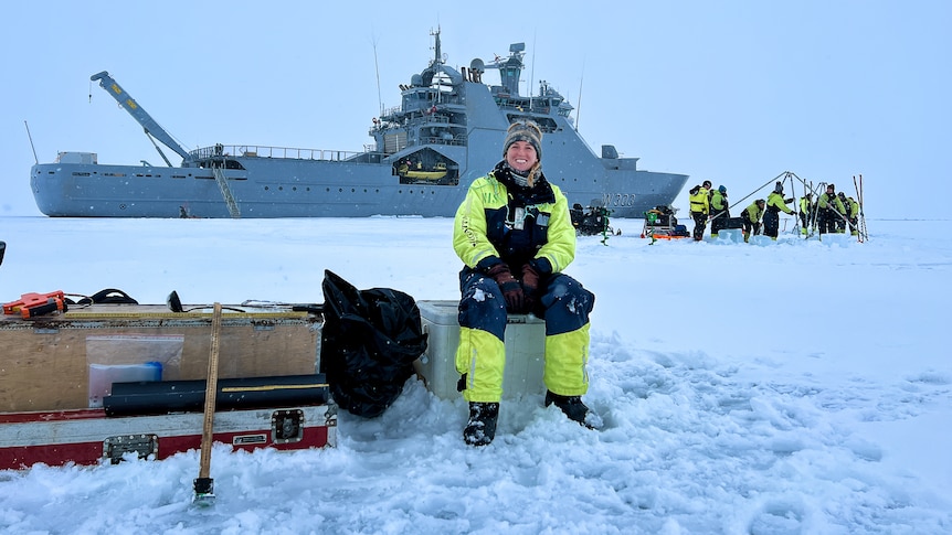 A smiling woman in cold weather gear sits in front of a large ship, surrounded by snow. 