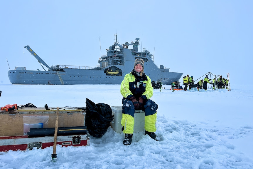 A smiling woman in cold weather gear sits in front of a large ship, surrounded by snow. 