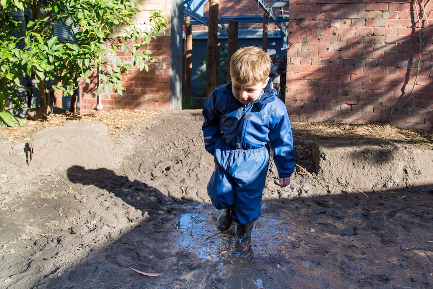 A little boy in blue wet weather gear and gumboots stomps in a mud puddle.