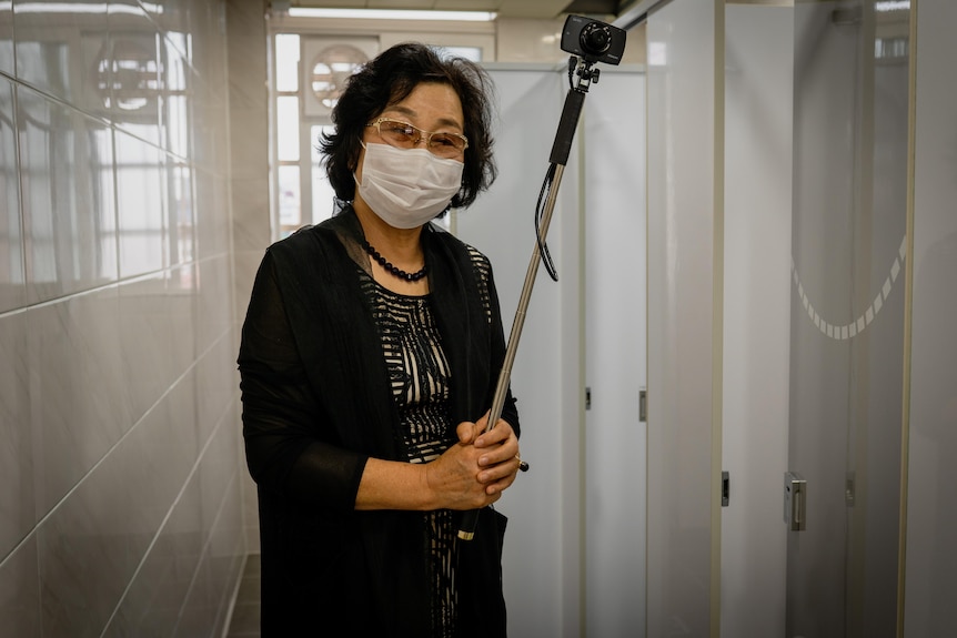 A Korean woman in a face mask holds a camera on a selfie stick in a public bathroom