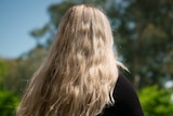A woman with long blonde hair stares out over a balcony with blurred greenery in the background, facing away and unidentified. 
