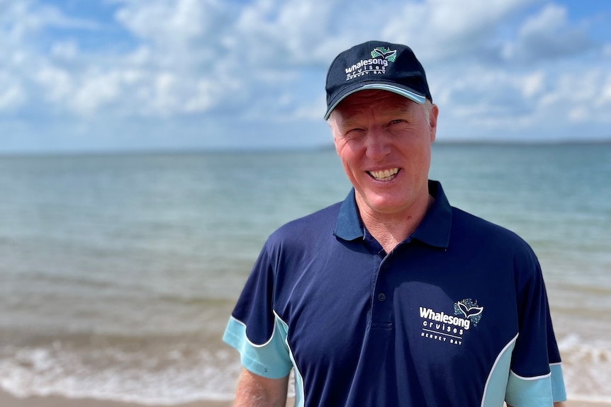 Man in a branded polo shirt and cap stands on the beach smiling 
