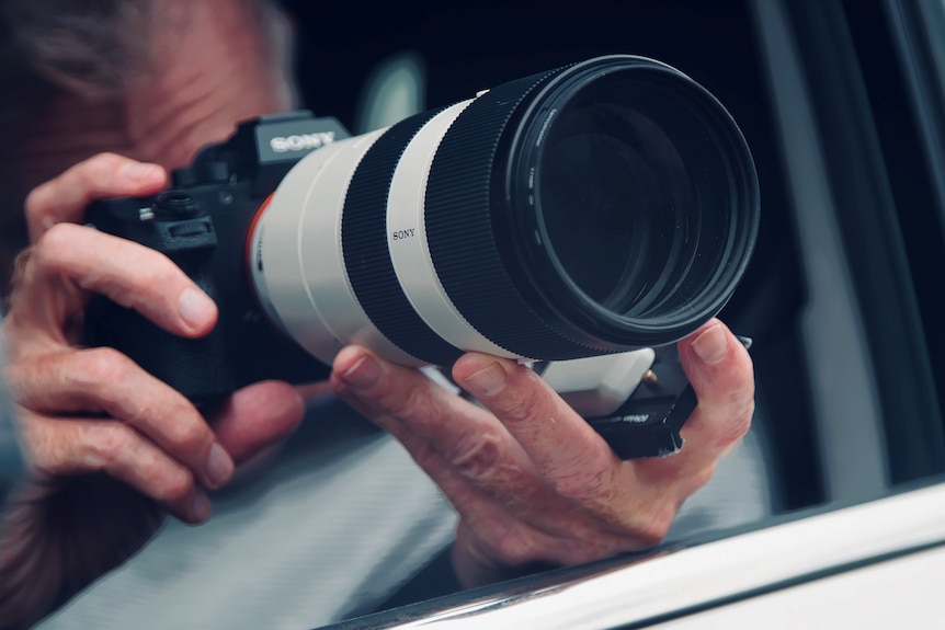 A man operates a camera with a long lens while sitting in a car.