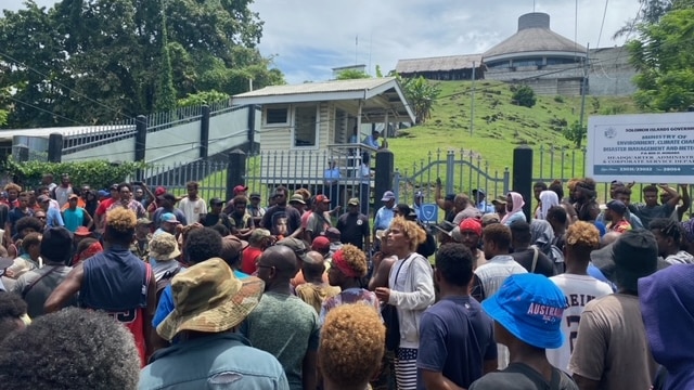 a crowd of people gather outside gates to Parliament House in Honiara