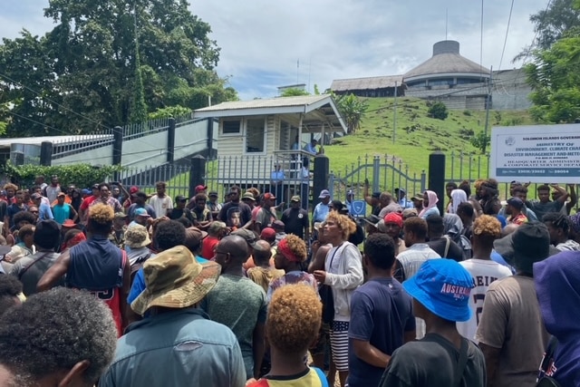 a crowd of people gather outside gates to Parliament House in Honiara