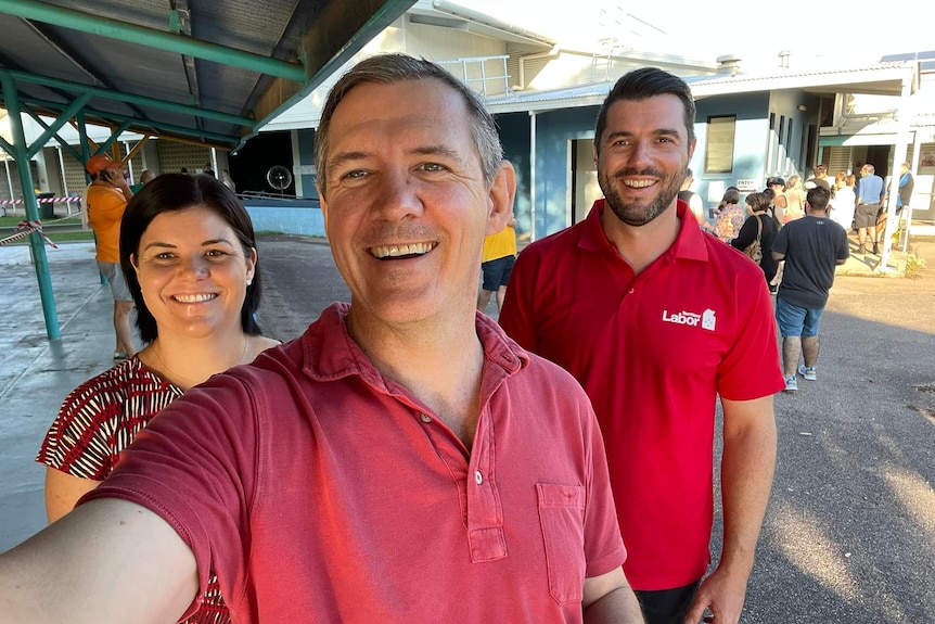 Three people smiling in a selfie, wearing red shirts for the Labor party. There's a line of voters at a primary school behind th