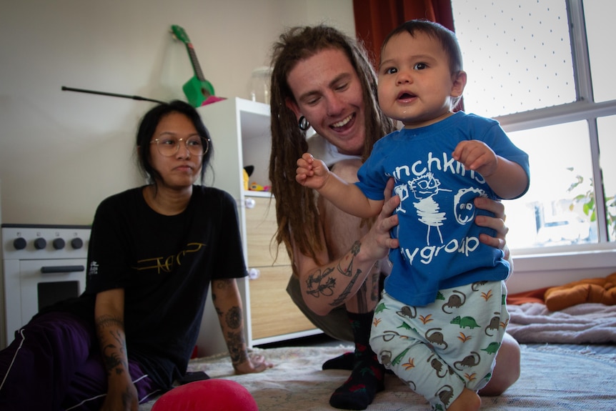 A man with dreadlocks holds an infant child, while a woman in black sits behind them