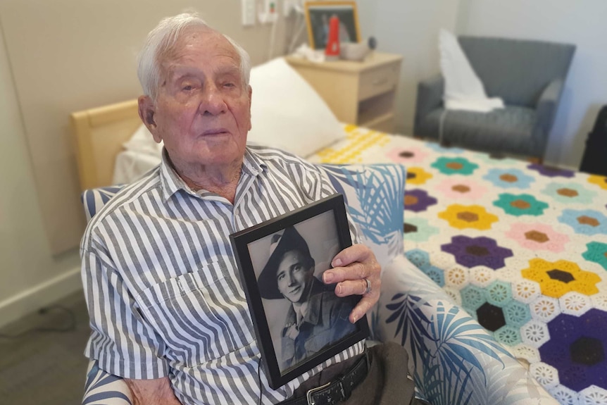 An older man sitting by a bed with a crocheted cover, holds a black and white photo of himself in Army uniform from 1941.